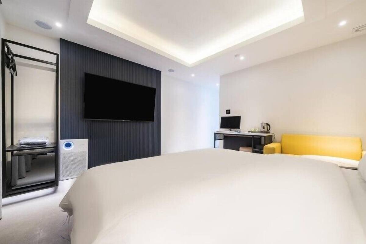 A modern hotel room with a large bed, a flat-screen TV mounted on the wall, a desk with a computer, and a yellow sofa.