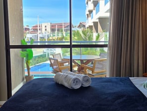 Habitación de hotel con cama, ventana con vista a una piscina y mobiliario exterior, además de una planta en maceta.