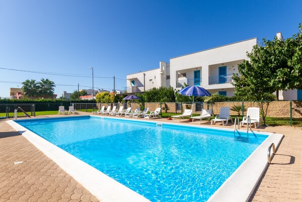 Una piscina rectangular con agua azul cristalina, rodeada de sillas de descanso y sombrillas blancas, con un edificio moderno al fondo.