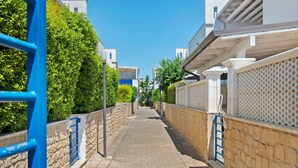 Un étroit sentier pavé, bordé de murs en pierre et de rampes bleues, mène à une série de bâtiments modernes aux façades blanches et aux touches de couleur bleue.