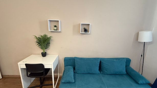 Een compacte, moderne kamer met een blauwgroene bank, een wit bureau met stoel, en twee kleine wandplanken met potplanten.