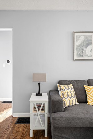 A room with a grey couch, a white side table with a lamp, a framed artwork on the wall, and a door.