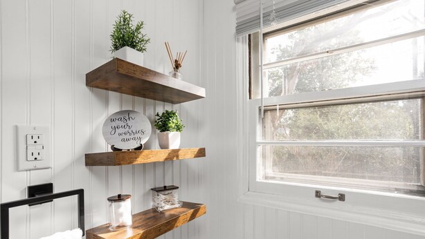 A bathroom with white paneled walls, a window with a view of trees, and wooden floating shelves with plants and a round mirror. - Pet-Friendly Broad Ripple Home W/ Fenced Backyard (Indianapolis)