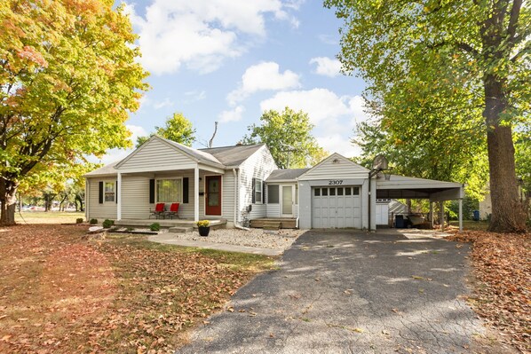 A two-story house with a covered driveway, a garage, and a porch with red chairs.