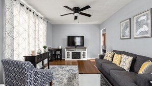A living room with a sofa, a patterned armchair, a coffee table, a TV, a fireplace, a ceiling fan, and artwork on the wall. - Pet-Friendly Broad Ripple Home W/ Fenced Backyard (Indianapolis)