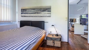 A modern hotel room with a bed, a painting of the ocean, a desk with a lamp, and a TV.