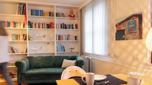 Un salon avec un canapé vert, une bibliothèque, une table à manger avec des assiettes et une tasse, et un panneau sur lequel on peut lire : « LIVRAISON DU PORT À LA TERRE ».