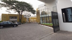 A modern building with a glass entrance, a paved driveway, and a parking area with cars. - The Cabin Hotel (Accra)
