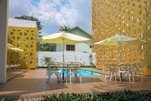 A poolside area with chairs and umbrellas, a pool, and a white building with a green roof. - The Cabin Hotel (Accra)