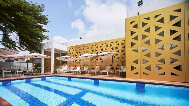 A swimming pool with lounge chairs and umbrellas, surrounded by a yellow patterned wall. - The Cabin Hotel (Accra)