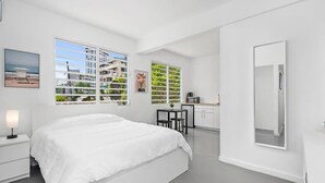 Un dormitorio moderno con una cama grande, una mesita de noche, un escritorio con una silla, un espejo y vistas al exterior a través de amplias ventanas.