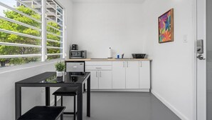 Cocina compacta con alacenas blancas, microondas y lavaplatos. Se ve una ventana con persianas, una mesita con una planta y un cuadro colorido en la pared.