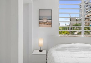 Un dormitorio con cama, una mesita de noche con lámpara, un cuadro enmarcado con escena de playa y una ventana que ofrece vistas de edificios y zonas verdes.