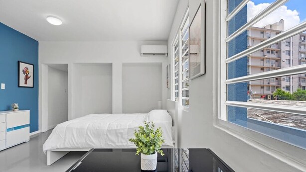 Un dormitorio moderno con una cama, una mesita de noche, una planta en maceta y un ventanal con vistas a la ciudad.