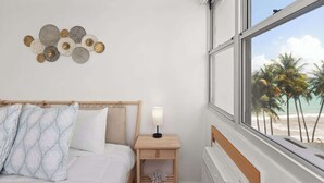 Een slaapkamer met een bed, een nachtkastje met een lamp, een kunstwerk aan de muur en een raam met uitzicht op het strand.