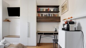 Een kleine kamer met een bed, een bureau, een stoel, een televisie en planken met boeken en decoratieve objecten.