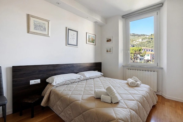 Un dormitorio con cama, una silla, una ventana con vistas y cuadros en la pared.