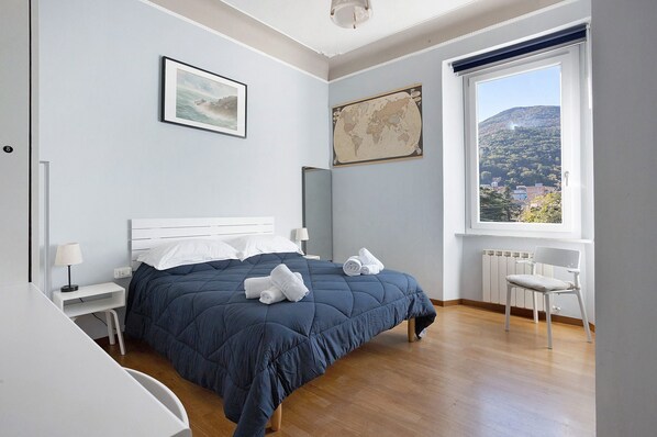 Un dormitorio con cama, una silla, un escritorio, una lámpara, un mapa en la pared y una ventana con vistas a una colina.
