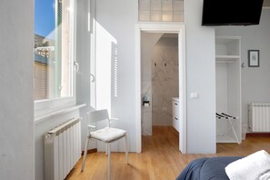 Un dormitorio con cama, una silla, un radiador, televisión y baño con ducha e inodoro.