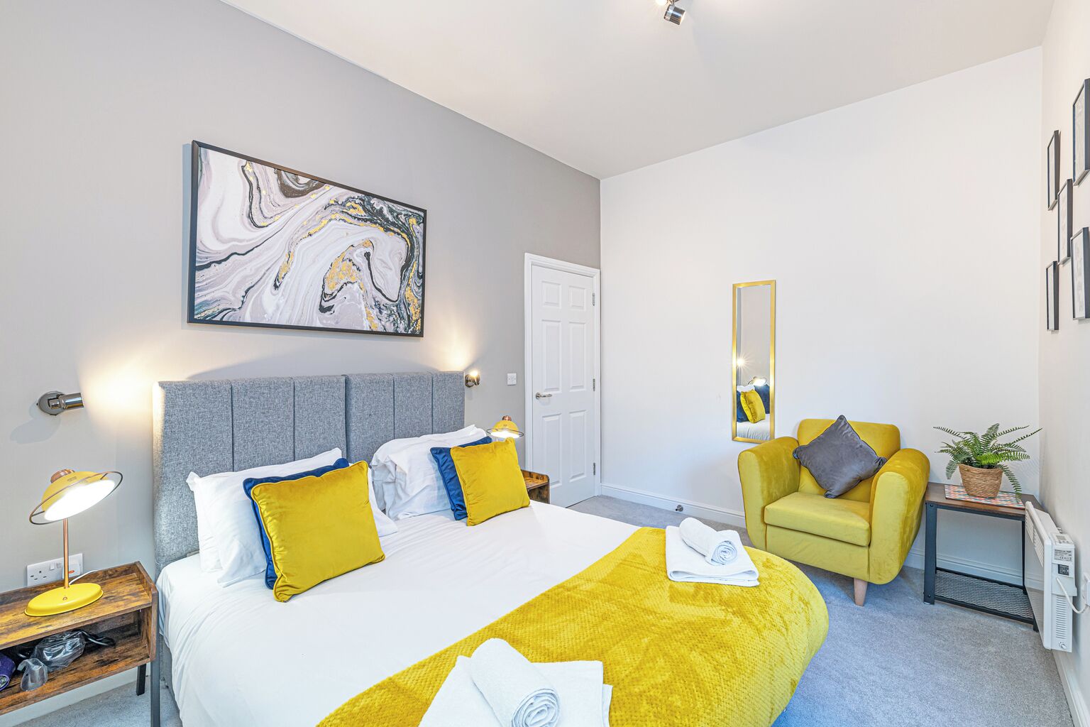 A modern hotel room with a large bed, a bedside table with a lamp, a chair, a mirror, and a painting on the wall.
