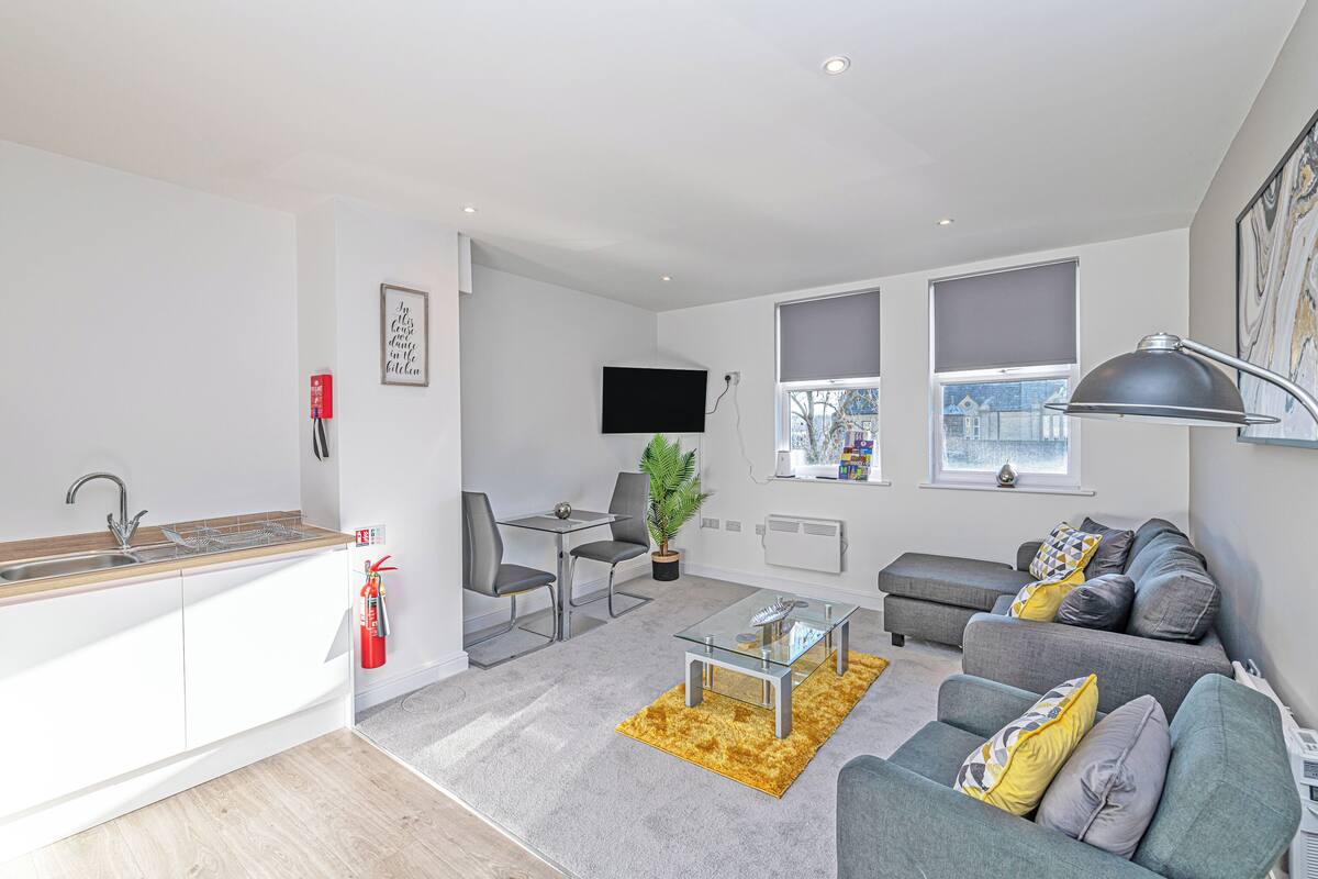 A modern living room with a grey sofa, a glass coffee table, and a small dining area with a table and chairs.