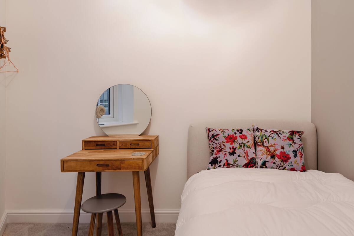 A bedroom with a wooden desk, a round mirror, a stool, and a bed with floral pillows.
