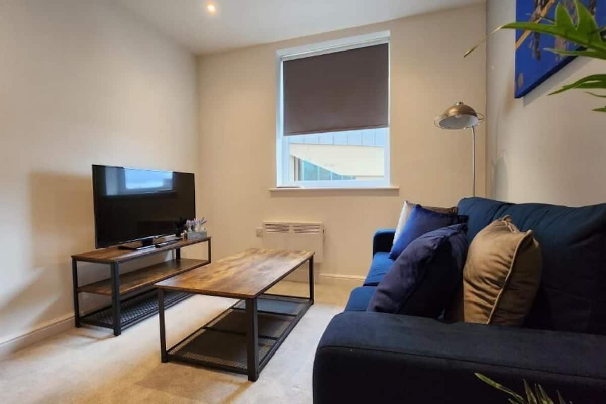 A living room with a flat-screen TV on a wooden coffee table, a dark sofa with cushions, a floor lamp, and a window with a blind.