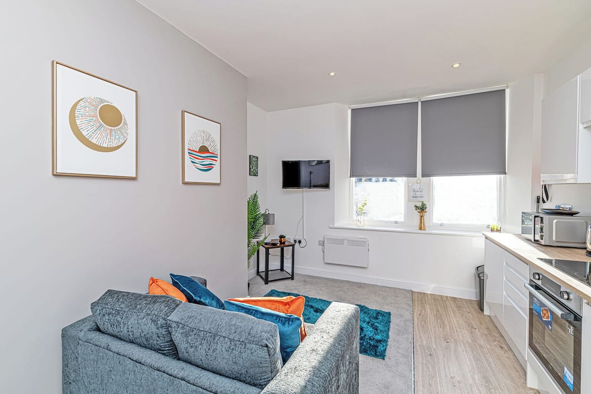 A modern apartment with a grey sofa, a kitchen with a microwave, and a TV on the wall.