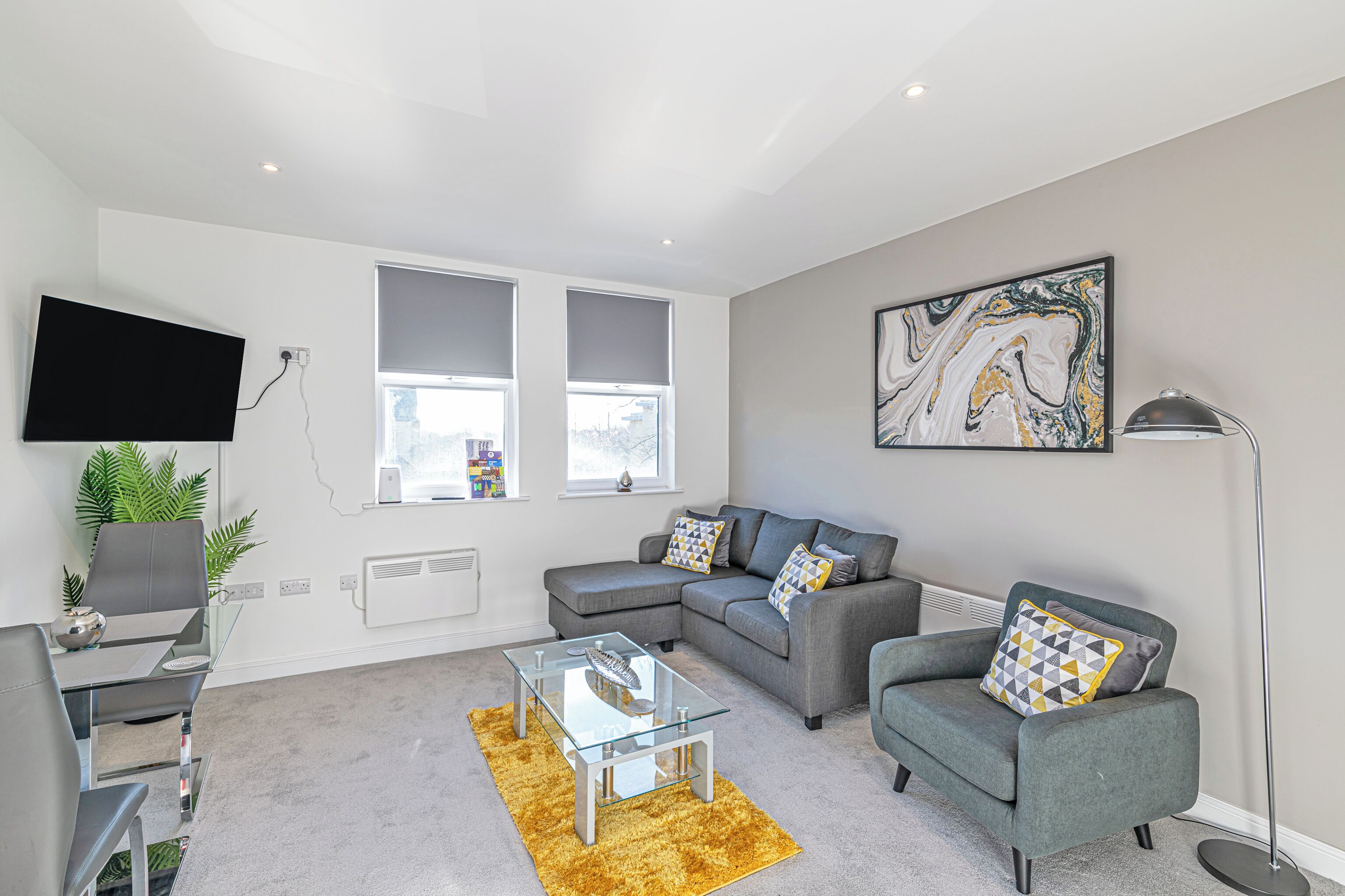 A modern living room with a grey sofa, a glass coffee table, a flat-screen TV mounted on the wall, and a floor lamp.