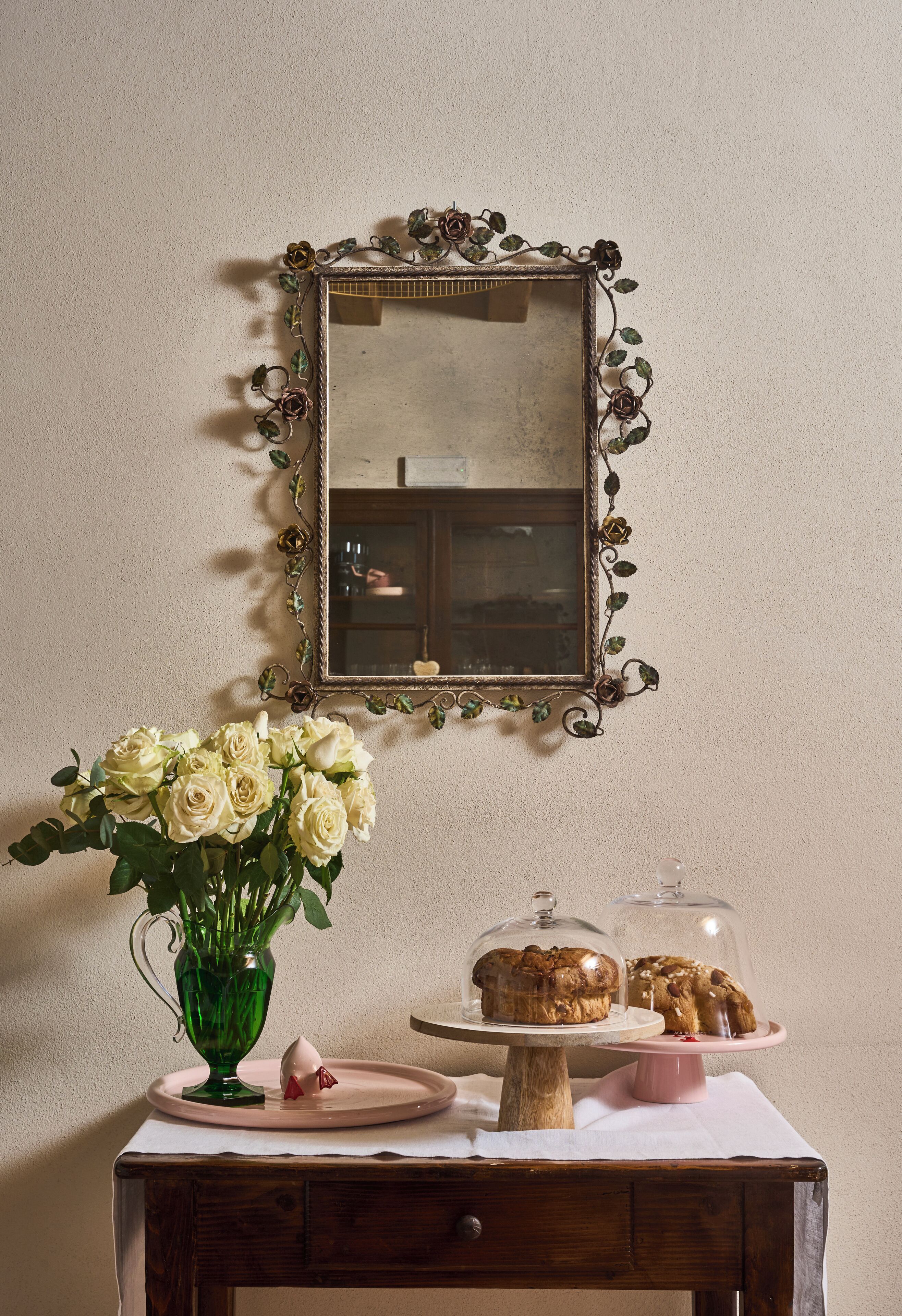 Un tavolo con un vaso di fiori, un vassoio con pasticcini e uno specchio con cornice decorativa.