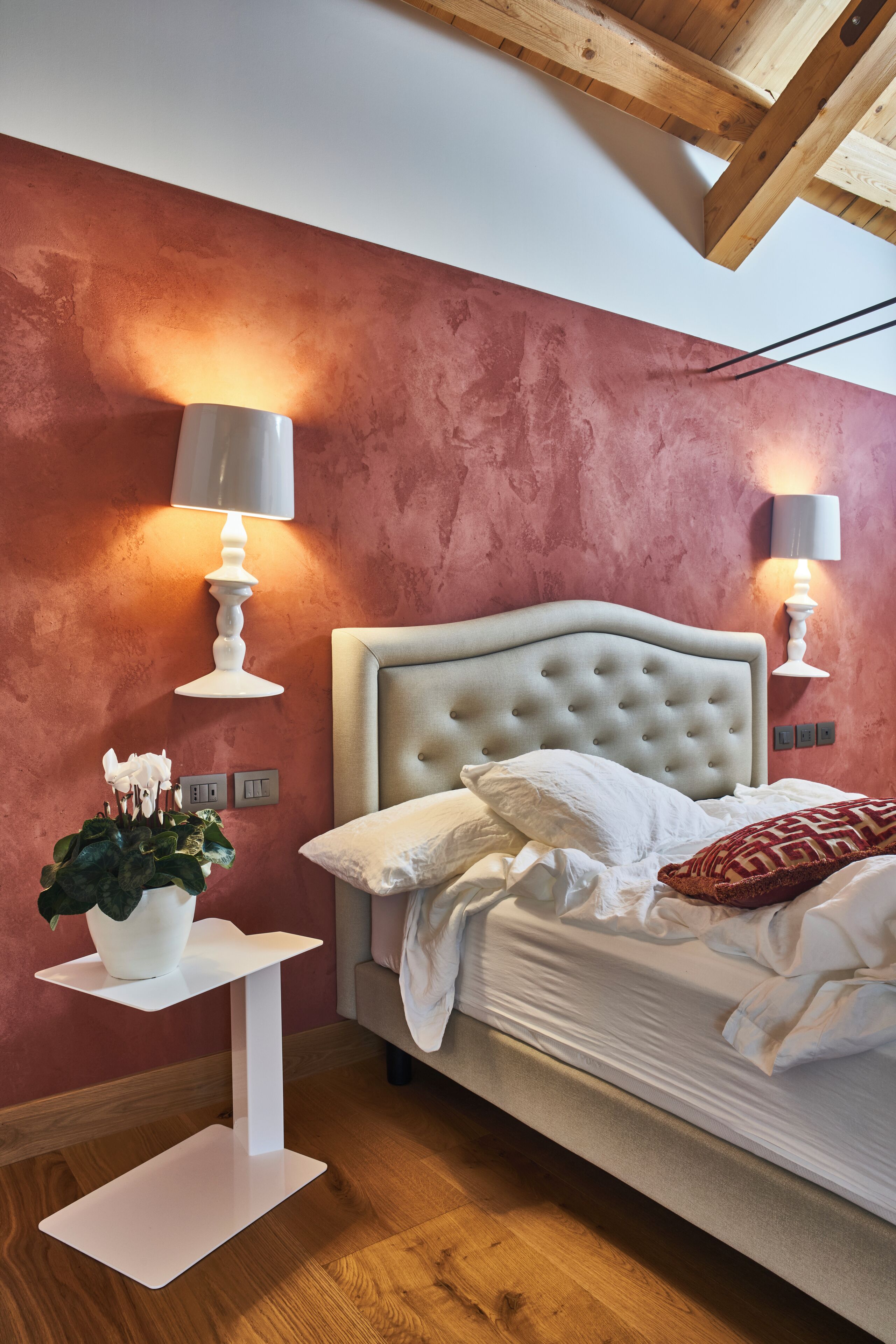 A bedroom with a tufted headboard, white bedding, two bedside lamps, a white bedside table with a plant, and a wooden ceiling.