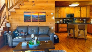 A cozy cabin interior with a wooden staircase, a leather sofa, a dining area, and a kitchen.
