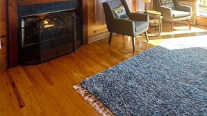 A cozy cabin interior with a fireplace, a flat-screen TV mounted above it, and two armchairs with a small table between them.