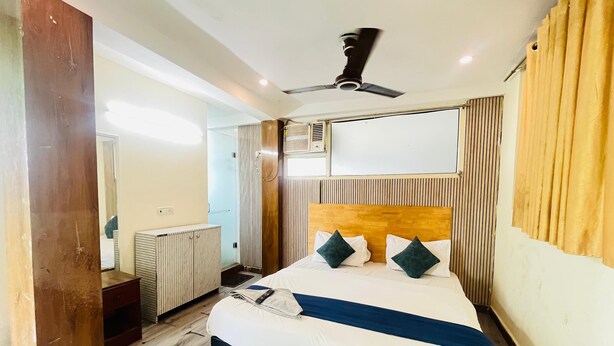 A hotel room with a bed, wooden headboard, a ceiling fan, a radiator, and a window with curtains. - Roomshala Hotel Green Valley (New Delhi)