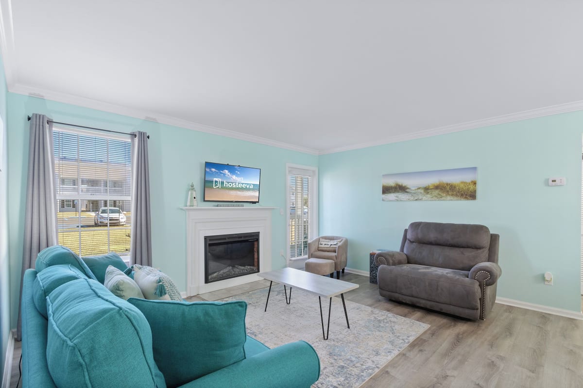 A living room with teal sofas, a fireplace, a television, and a view of the street.