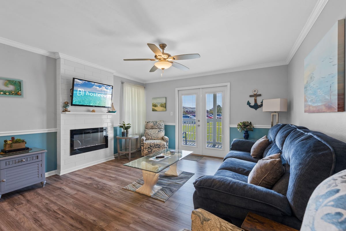 A living room with a fireplace, a television, a ceiling fan, and a large window.