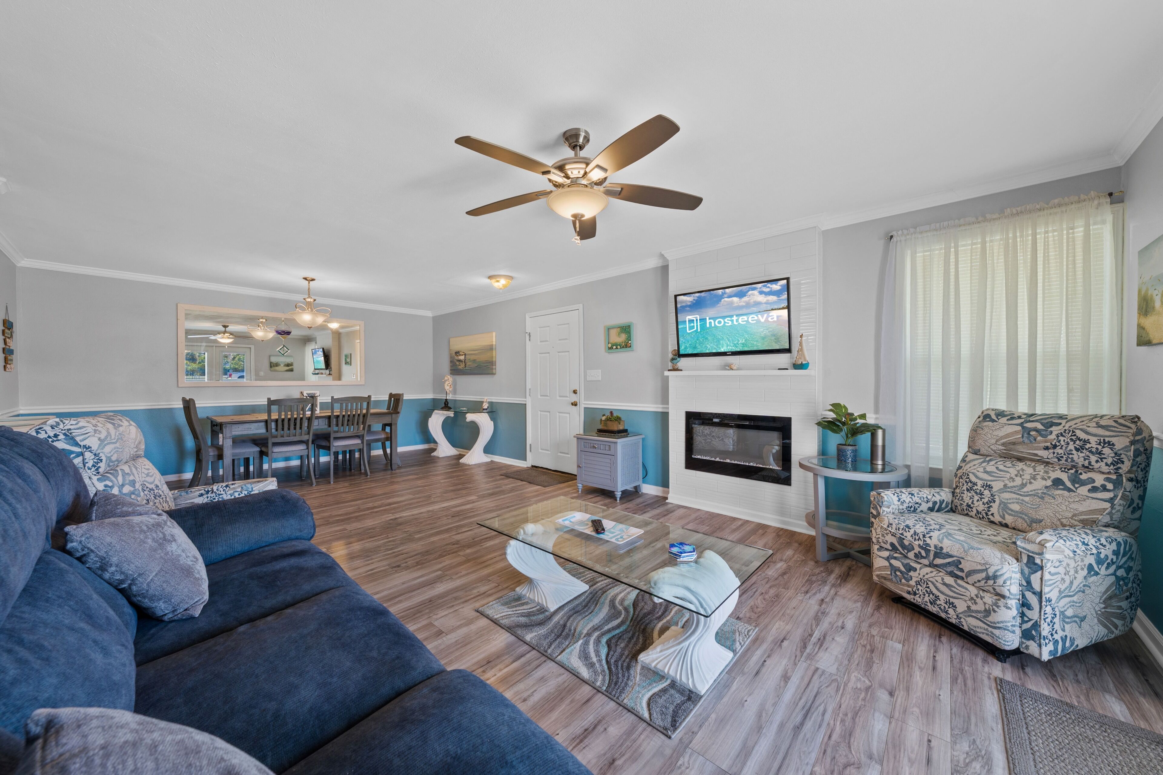 A living room with a sofa, armchair, glass coffee table, ceiling fan, TV, fireplace, and dining area.