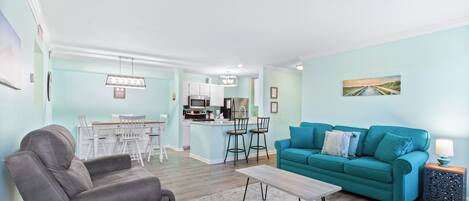 Una sala de estar con un sofá color turquesa, un sillón gris y una mesa de centro. Se observa una cocina al fondo con armarios blancos y un comedor con sillas blancas.
