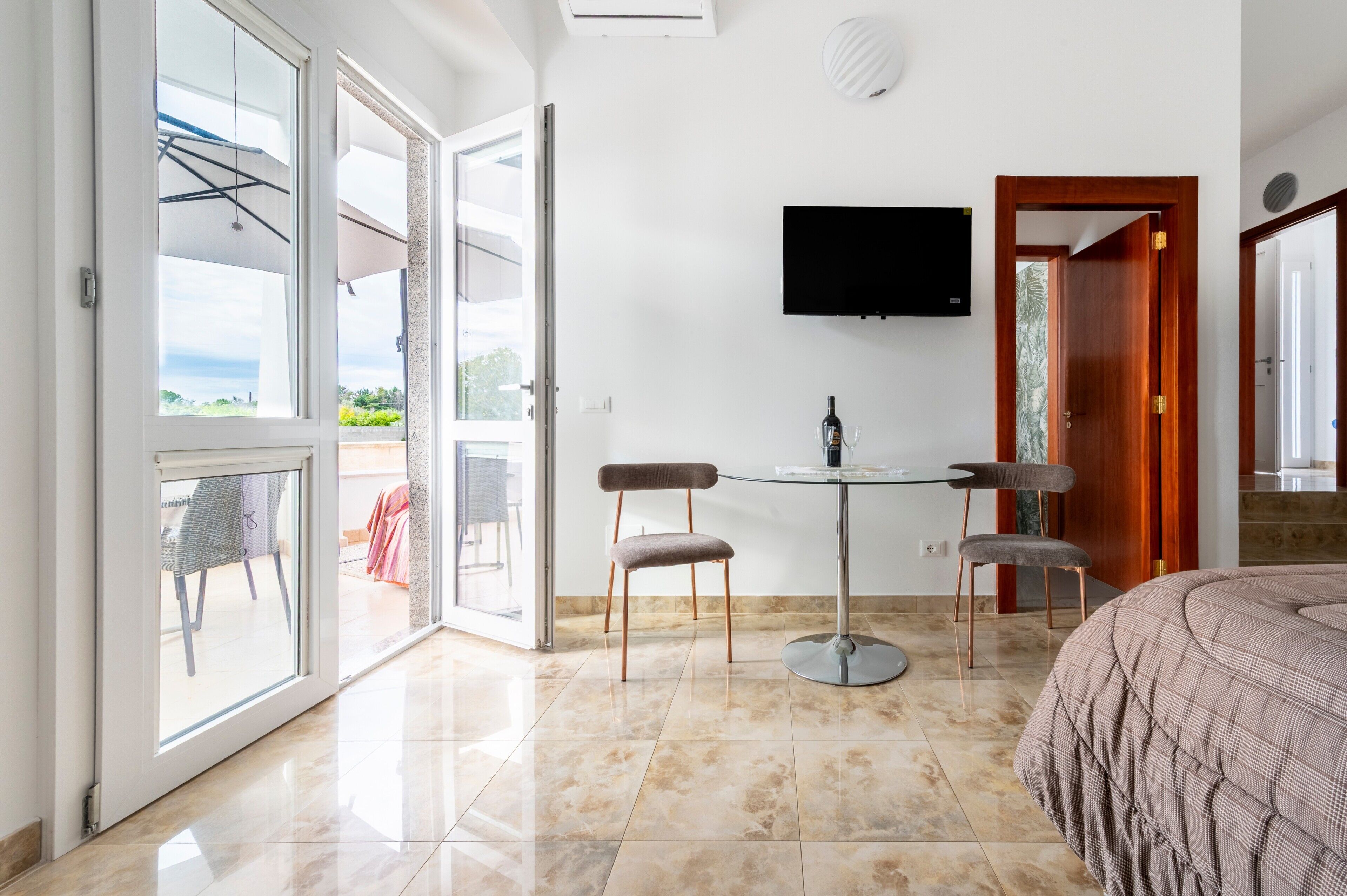 Habitación de hotel con cama, televisor montado en la pared, mesa con sillas y puerta corrediza de vidrio que da a un área exterior.
