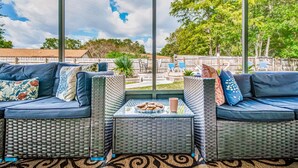 A covered outdoor seating area with wicker furniture and cushions, overlooking a backyard with a pool and lounge chairs.