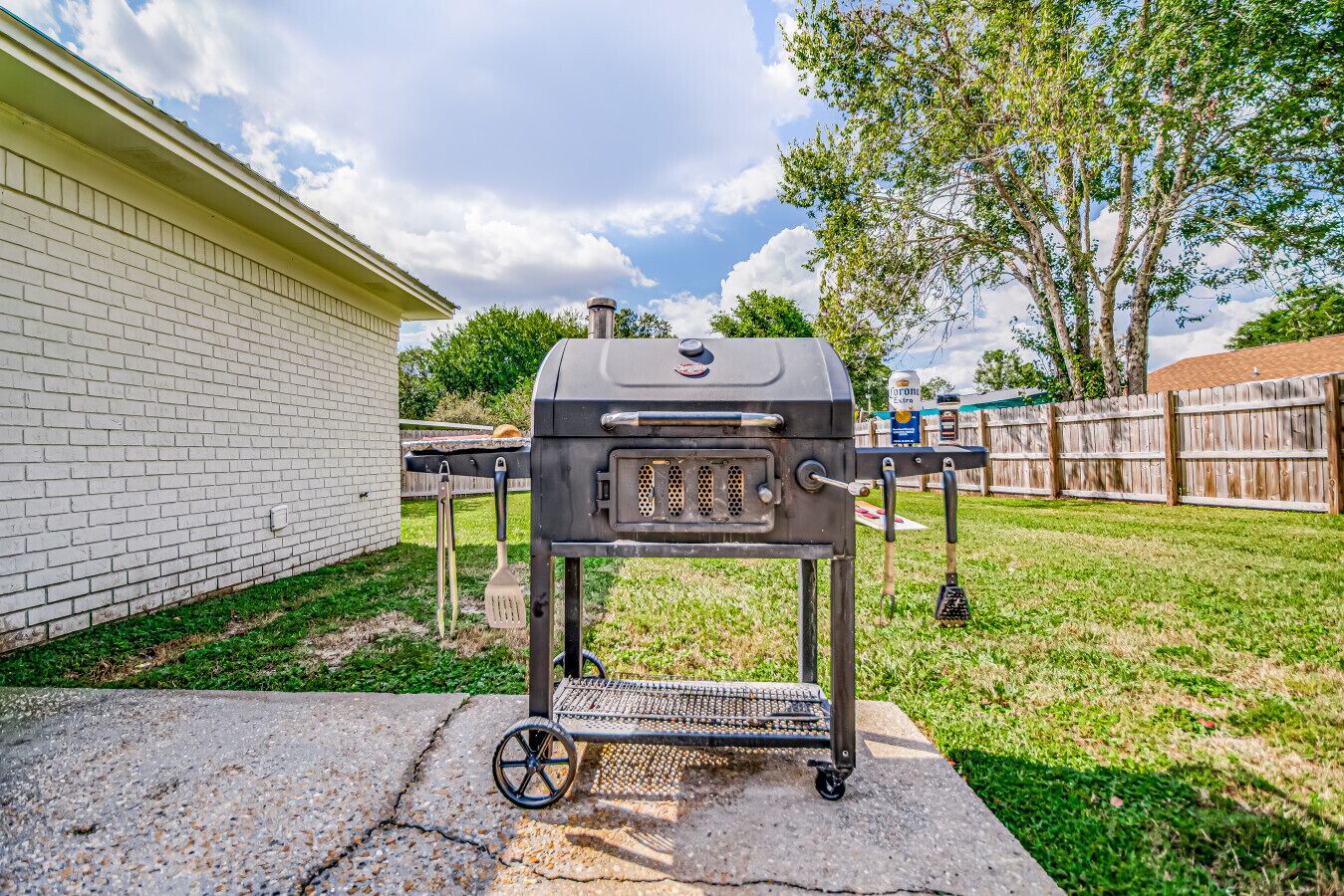 Een draagbare barbecue met wielen, geplaatst op een betonnen terras naast een huis met witte bakstenen.