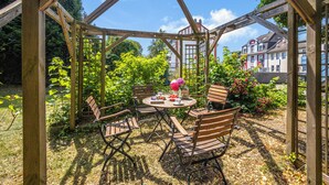 Une structure de pergola en bois, avec une table ronde et des chaises, entourée d’une végétation luxuriante et offrant une vue sur un immeuble d’habitation.