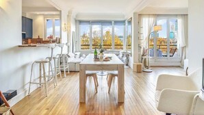 Un soggiorno moderno con un tavolo da pranzo in legno, sgabelli da bar bianchi e un'ampia finestra che si affaccia su alberi autunnali e edifici.