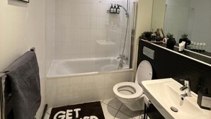 Un baño moderno con bañera, inodoro y lavamanos. Hay una alfombra blanco y negro en el suelo con el texto "GET NAKED".