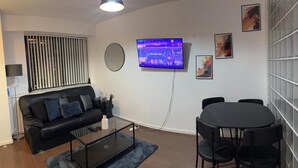 Una sala moderna con un sofá de cuero negro, una mesa de centro de vidrio, una mesa redonda con cuatro sillas, un televisor montado en la pared y un ventanal con persianas.