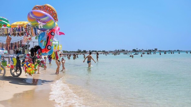 A beach with clear water, people swimming, and a vendor stand with colorful items.