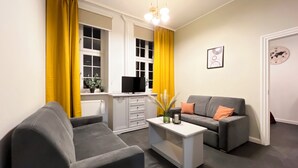 Ein modernes Wohnzimmer mit zwei grauen Sofas, einem weißen Couchtisch und einem Fernseher auf einer Kommode. Gelbe Vorhänge und eine Wanduhr mit Weltkarten sind sichtbar.