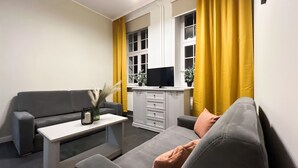 Ein modernes Wohnzimmer mit einem grauen Sofa, einem weißen Couchtisch und einem Fernseher auf einem Schrank. Gelbe Vorhänge sind sichtbar.