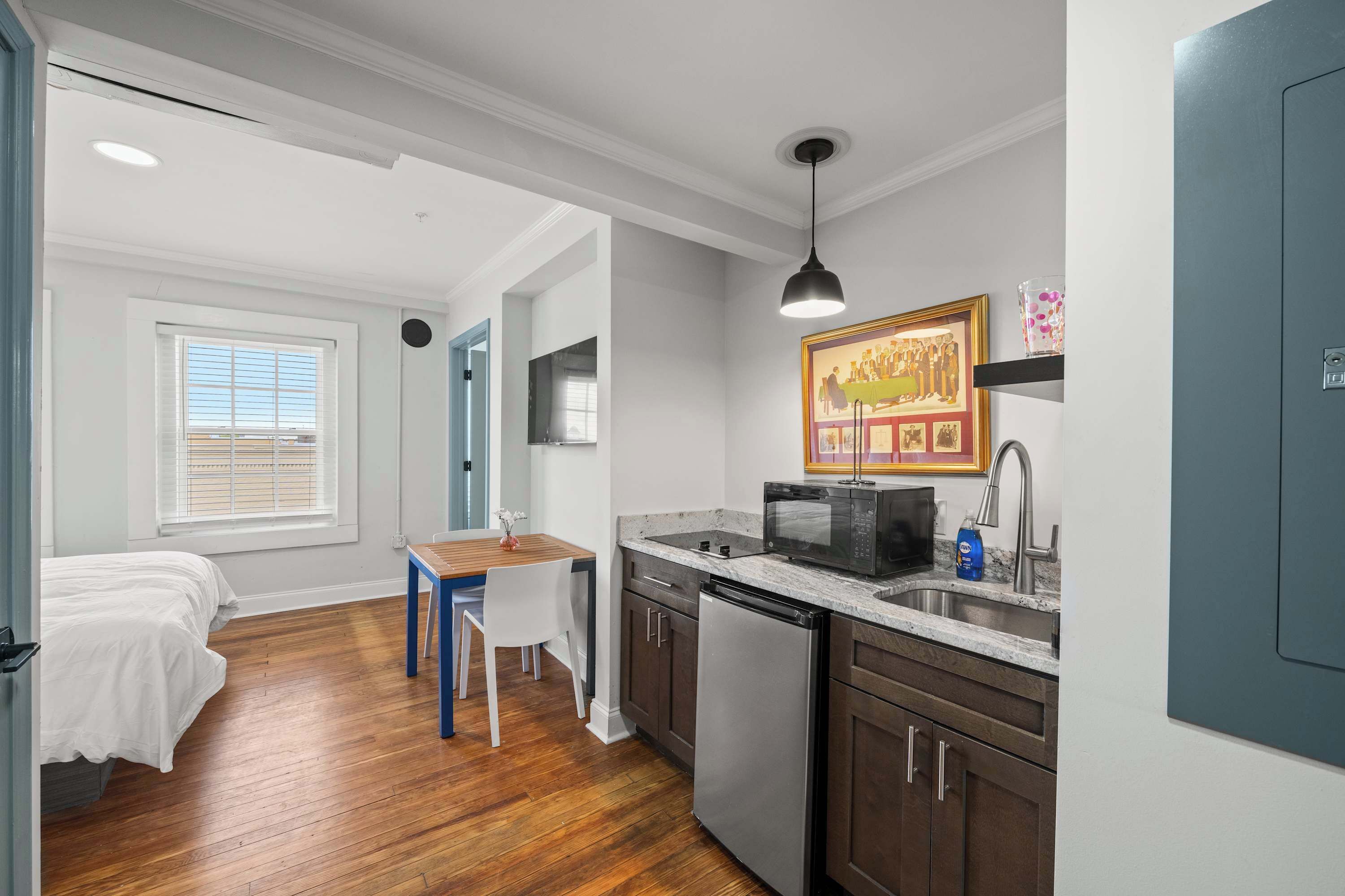 A compact kitchen with a microwave, sink, and dishwasher. There is a dining table with chairs, and a bed is visible in the adjacent room.