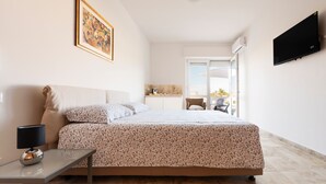Une chambre à coucher avec un lit, une table de chevet avec une lampe, un téléviseur fixé au mur et un coin cuisine visible à travers une porte ouverte.
